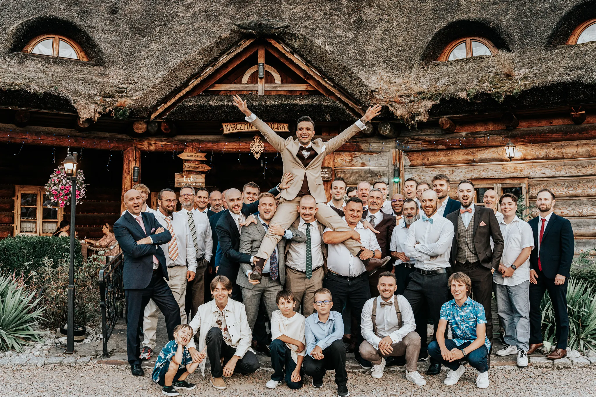 radosne zdjecie slubne mezczyzn na weselu unoszacyhc mlodego pana w bezowym garniturze wykonane przed sala weselna mlynowka